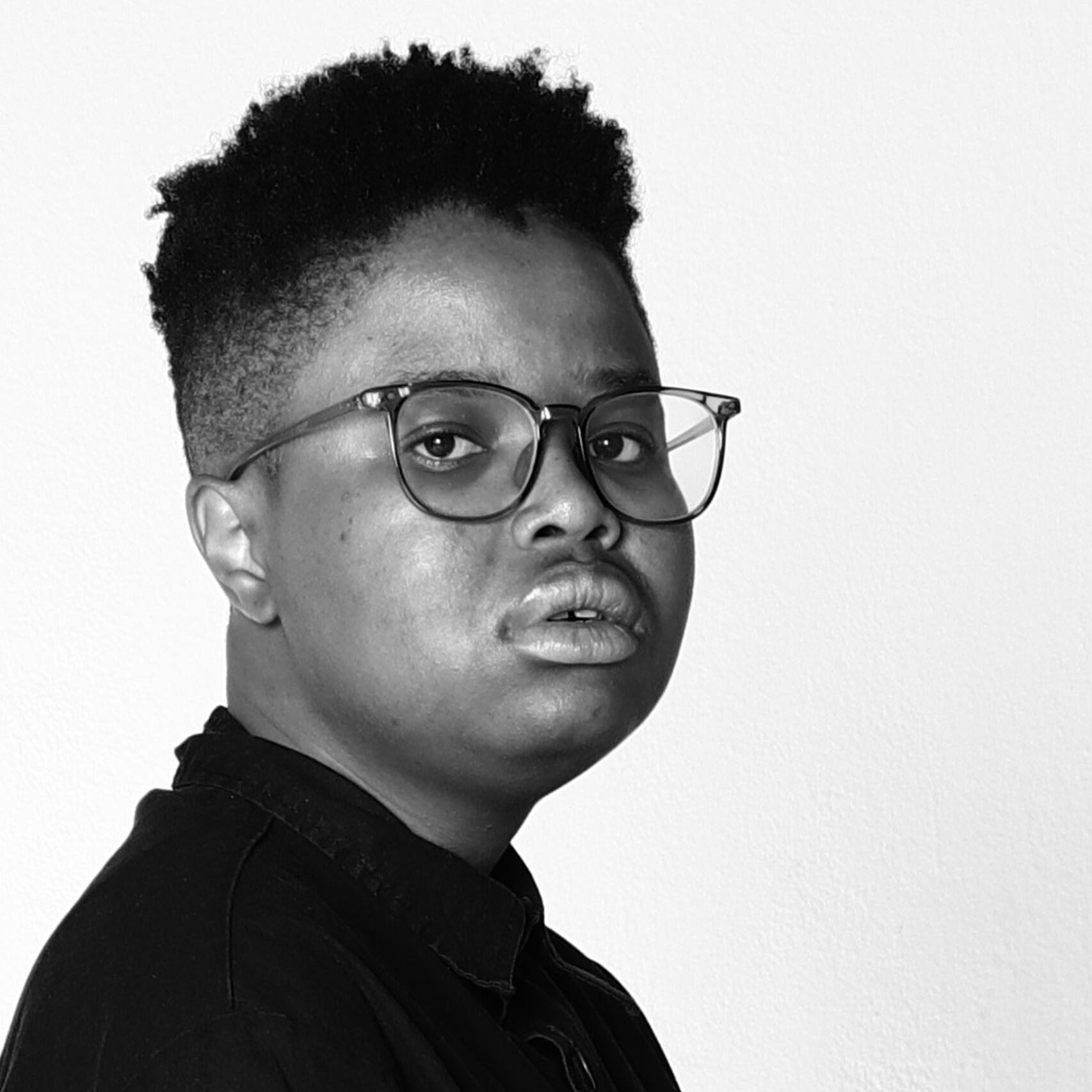 A black and white close up photo of Carbon. They are dressed in a black shirt against a white background with glasses and a short fade haircut. They are three quarter turned towards the camera with a serious expression on their face.