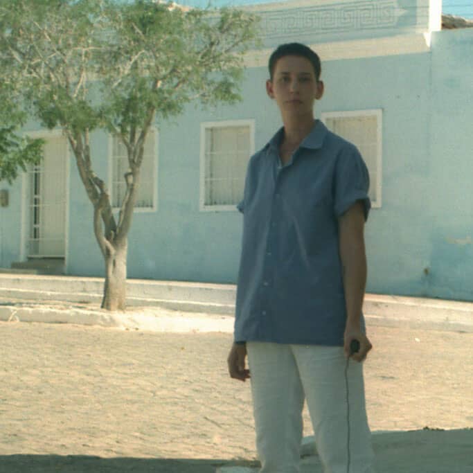Image Description: Photo of jialu pombo on an empty cobblestone street. They have white skin, short brown hair, and a thin body. They stand in the distance, look at the camera and hold the remote photo trigger, they wear white pants and a blue shirt. In t