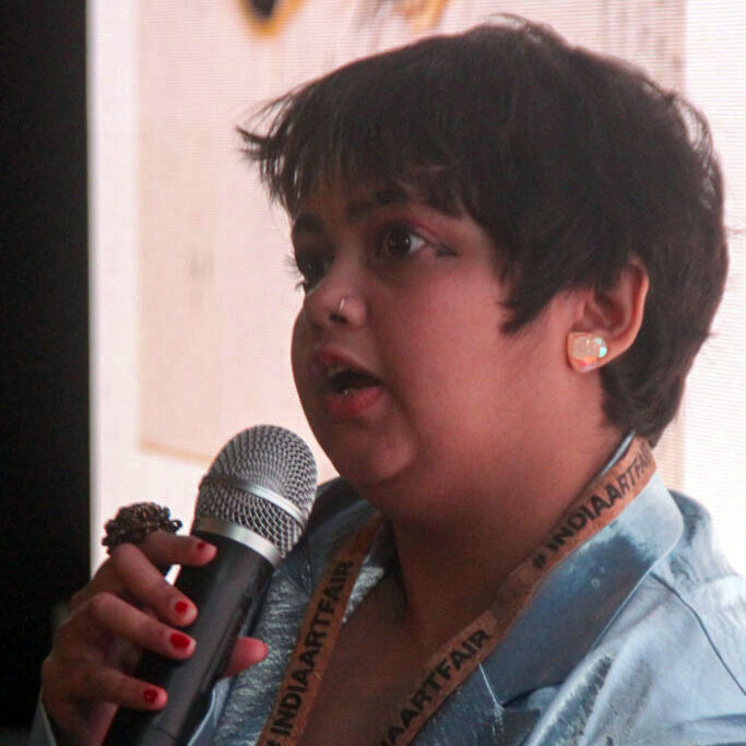 Image Description: Nu can be seen holding a microphone and talking. They have short brown hair and are wearing a turquoise coat.