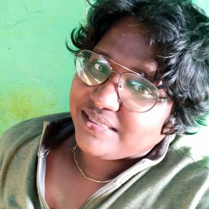 Image Description: A close-up photo of Vino. They are wearing a green t-shirt against a green background. They have short black hair and are wearing glasses. They are turned towards the camera with a smile on their face.